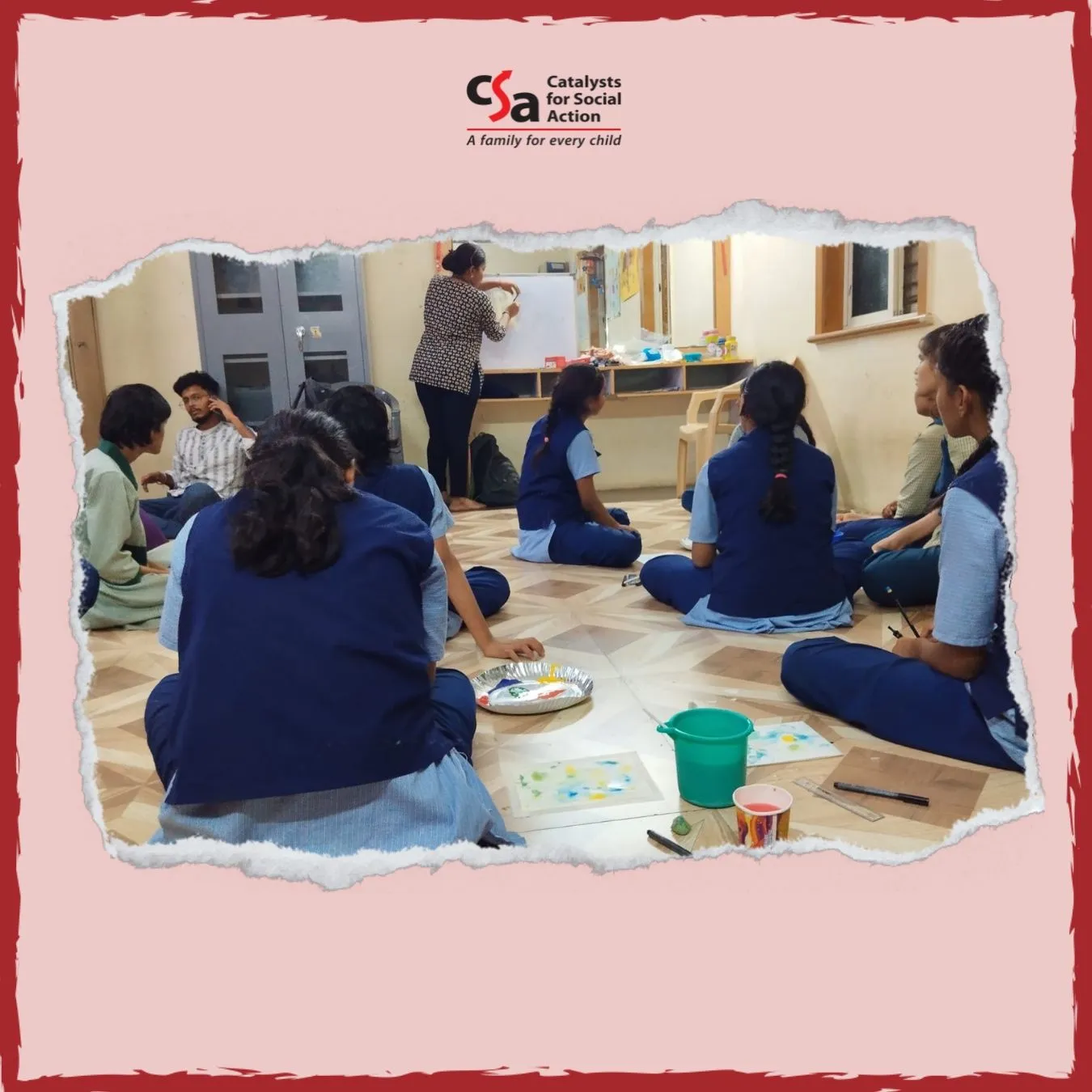A group of school-age girls in uniforms are seated on the floor in a classroom, engaging in an art activity. They are using paint, brushes, and drawing sheets placed on the floor. A woman stands at the front drawing on a whiteboard, guiding the session. The setting has a warm, inclusive atmosphere. The image is framed with a torn-paper effect and features the logo of "Catalysts for Social Action" with the tagline "A family for every child" at the top.