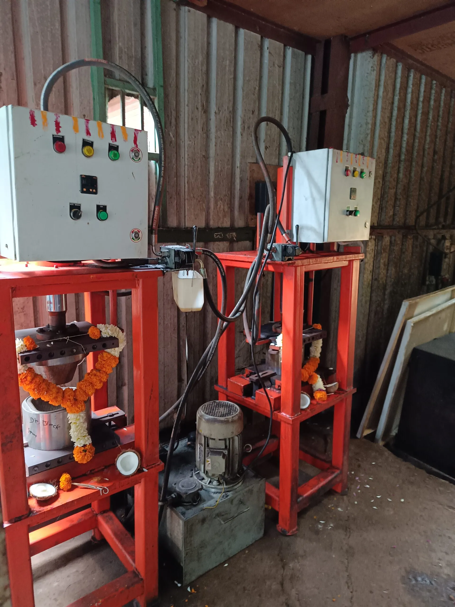 Two industrial hydraulic press machines with red frames and white control panels are positioned side by side inside a corrugated metal shed. Each machine has a garland of marigold and white flowers draped over it, indicating they have been recently inaugurated or are part of a ceremonial event. Wires and hoses are connected to the machines, and a metal motor unit is visible on the ground between them. The environment appears to be a workshop or small-scale manufacturing unit.